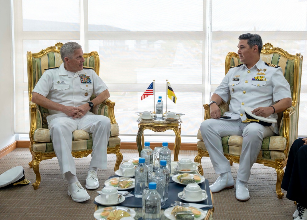 Two naval officers in white dress uniforms are seated in ornate gold-framed chairs engaged in a bilateral meeting. Between them on a small decorative table are two small flags — one American and one appearing to be from Brunei — indicating an official diplomatic or military exchange. A refreshment table with water bottles, cups, and food is positioned in the foreground.