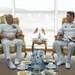 Two naval officers in white dress uniforms are seated in ornate gold-framed chairs engaged in a bilateral meeting. Between them on a small decorative table are two small flags — one American and one appearing to be from Brunei — indicating an official diplomatic or military exchange. A refreshment table with water bottles, cups, and food is positioned in the foreground.