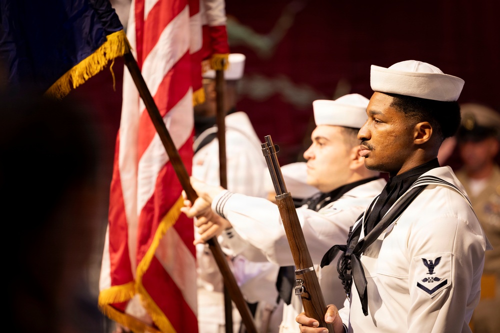 A U.S. Navy color guard is participating in a formal military ceremony. Sailors in dress white uniforms are presenting the American flag and carrying ceremonial rifles. The foreground sailor displays a Petty Officer rating badge on his left sleeve.