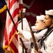 A U.S. Navy color guard is participating in a formal military ceremony. Sailors in dress white uniforms are presenting the American flag and carrying ceremonial rifles. The foreground sailor displays a Petty Officer rating badge on his left sleeve.