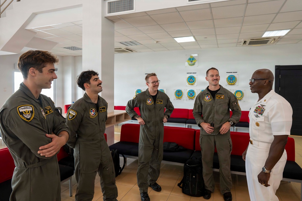 Five military personnel are engaged in a casual conversation in what appears to be a military facility briefing or common room. Four individuals are wearing olive/sage green flight suits with squadron patches, while one senior officer on the right is dressed in a white Navy dress uniform with multiple ribbons and medals. The atmosphere appears relaxed and collegial, with several individuals smiling or laughing.