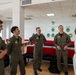 Five military personnel are engaged in a casual conversation in what appears to be a military facility briefing or common room. Four individuals are wearing olive/sage green flight suits with squadron patches, while one senior officer on the right is dressed in a white Navy dress uniform with multiple ribbons and medals. The atmosphere appears relaxed and collegial, with several individuals smiling or laughing.