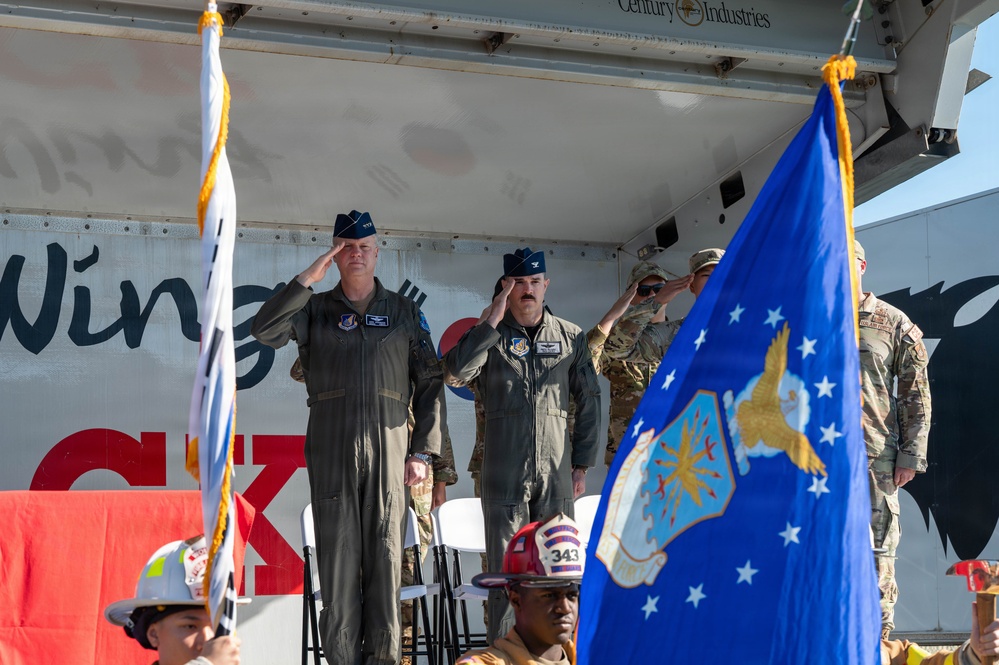 A military ceremony is taking place on an outdoor stage, with U.S. Air Force personnel in flight suits and service caps rendering salutes. A blue Air Force unit flag with a gold eagle emblem is prominently displayed in the foreground, alongside what appears to be a South Korean flag. Firefighters wearing helmets, including one marked '343', are visible in the foreground.