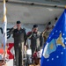 A military ceremony is taking place on an outdoor stage, with U.S. Air Force personnel in flight suits and service caps rendering salutes. A blue Air Force unit flag with a gold eagle emblem is prominently displayed in the foreground, alongside what appears to be a South Korean flag. Firefighters wearing helmets, including one marked '343', are visible in the foreground.