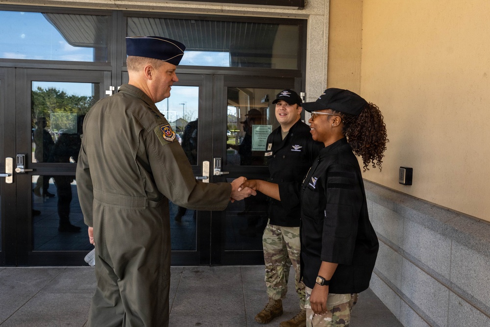A senior U.S. Air Force officer in an olive green flight suit and service cap is shaking hands with a person wearing a black uniform and cap outside a military building. A second individual in a black uniform and camouflage pants stands nearby observing the greeting. The setting appears to be the entrance of a military facility.