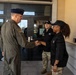 A senior U.S. Air Force officer in an olive green flight suit and service cap is shaking hands with a person wearing a black uniform and cap outside a military building. A second individual in a black uniform and camouflage pants stands nearby observing the greeting. The setting appears to be the entrance of a military facility.