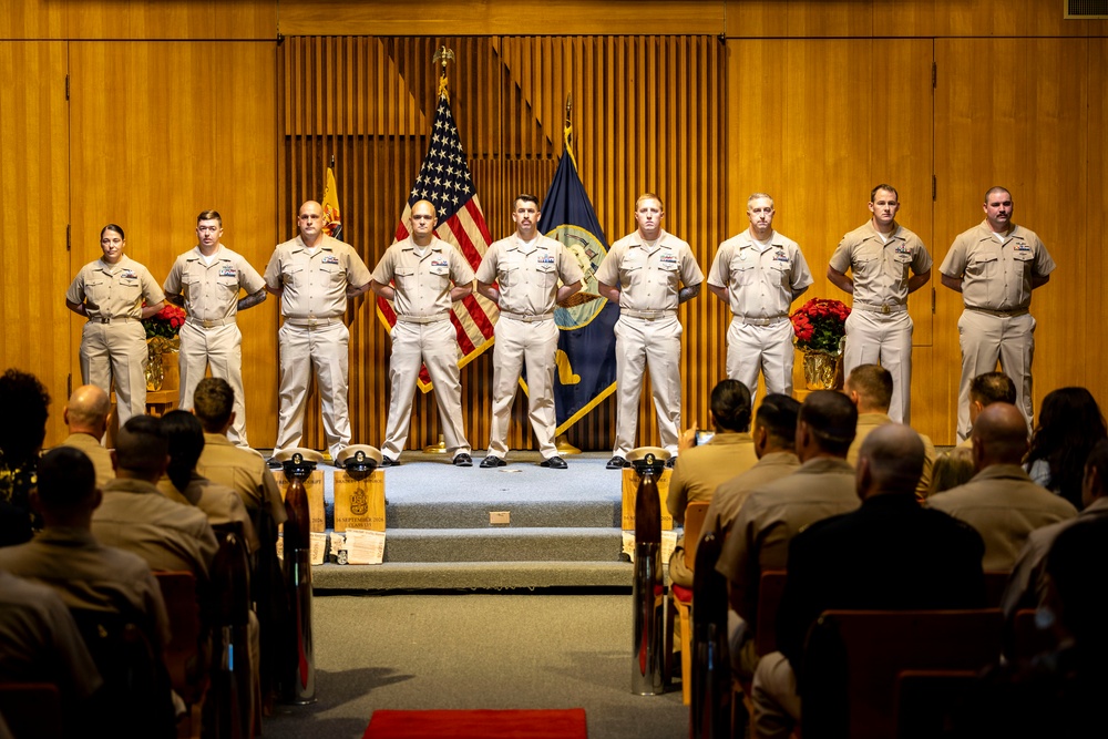 A formal U.S. Navy military ceremony is taking place in an auditorium or ceremonial hall. Nine uniformed Navy personnel stand at attention on a raised stage, flanked by the American flag and a Navy flag, while an audience of seated military personnel observes from the front. The setting includes decorative floral arrangements and a wooden podium or ceremonial box at the base of the stage.