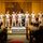 A formal U.S. Navy military ceremony is taking place in an auditorium or ceremonial hall. Nine uniformed Navy personnel stand at attention on a raised stage, flanked by the American flag and a Navy flag, while an audience of seated military personnel observes from the front. The setting includes decorative floral arrangements and a wooden podium or ceremonial box at the base of the stage.