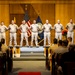 A formal U.S. Navy military ceremony is taking place in an auditorium or ceremonial hall. Nine uniformed Navy personnel stand at attention on a raised stage, flanked by the American flag and a Navy flag, while an audience of seated military personnel observes from the front. The setting includes decorative floral arrangements and a wooden podium or ceremonial box at the base of the stage.