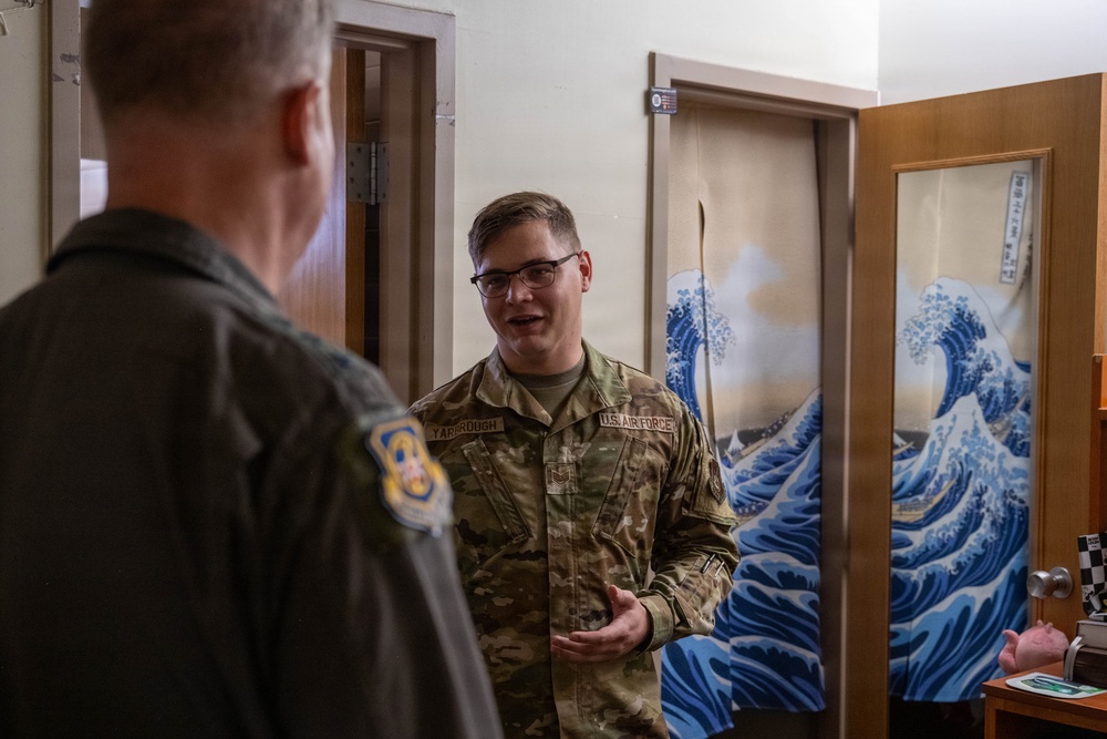 Two U.S. Air Force personnel are engaged in conversation inside what appears to be a dormitory or barracks room. The airman facing the camera wears an OCP (Operational Camouflage Pattern) uniform with a name tape reading 'YARBOROUGH' and 'U.S. AIR FORCE' tape. A decorative Japanese-style wave curtain, reminiscent of Hokusai's 'The Great Wave,' is visible on a door in the background.