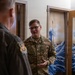 Two U.S. Air Force personnel are engaged in conversation inside what appears to be a dormitory or barracks room. The airman facing the camera wears an OCP (Operational Camouflage Pattern) uniform with a name tape reading 'YARBOROUGH' and 'U.S. AIR FORCE' tape. A decorative Japanese-style wave curtain, reminiscent of Hokusai's 'The Great Wave,' is visible on a door in the background.
