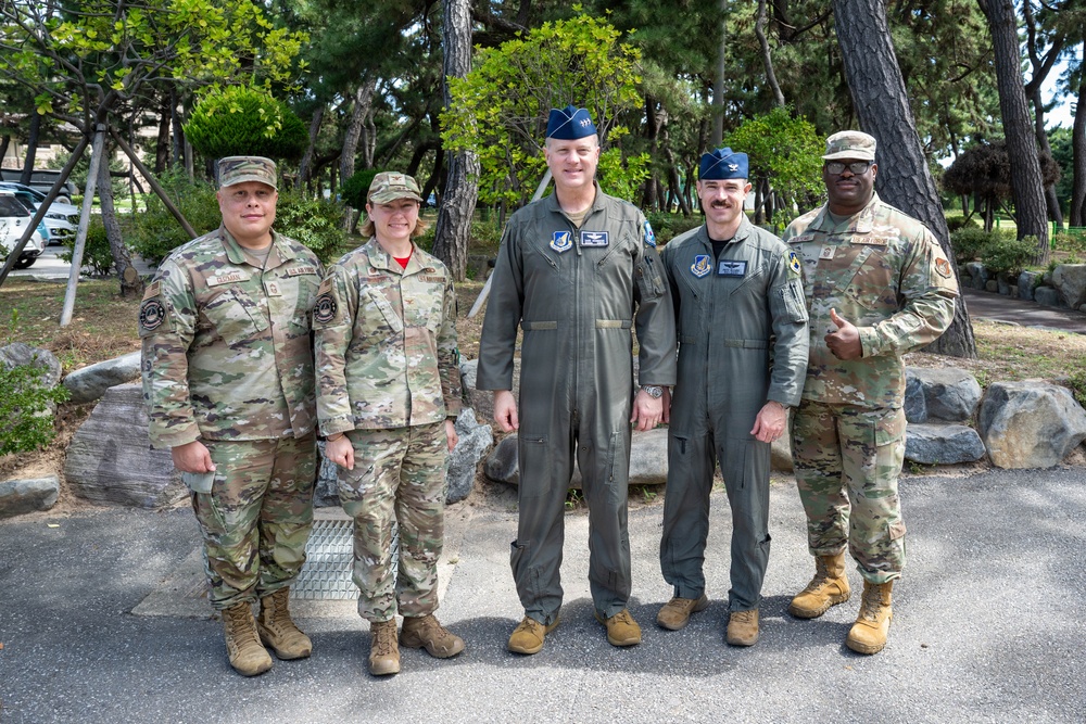 Five U.S. military personnel pose for a group photo outdoors on what appears to be a military installation surrounded by trees. Three individuals wear Army Combat Uniforms (ACU/OCP camouflage pattern) while two wear Air Force flight suits with blue garrison caps. The setting appears to be a well-maintained base environment with landscaped rocks and pine trees in the background.