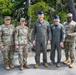 Five U.S. military personnel pose for a group photo outdoors on what appears to be a military installation surrounded by trees. Three individuals wear Army Combat Uniforms (ACU/OCP camouflage pattern) while two wear Air Force flight suits with blue garrison caps. The setting appears to be a well-maintained base environment with landscaped rocks and pine trees in the background.