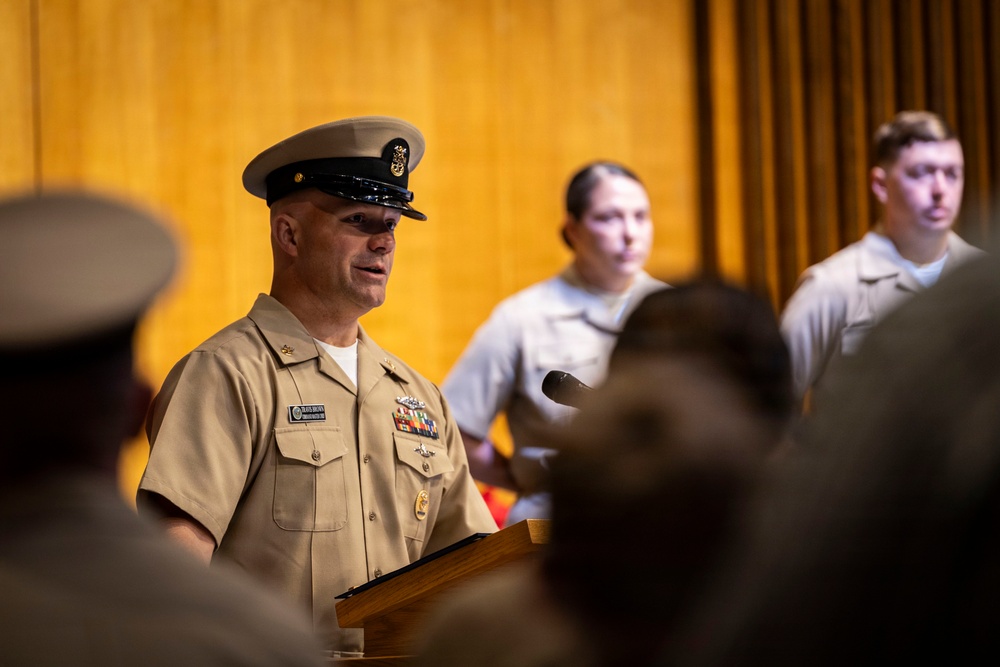 A senior U.S. Navy Chief Petty Officer in Service Khaki uniform is speaking at a podium during what appears to be a formal military ceremony or event. Multiple service members in lighter utility uniforms are visible in the background. The setting appears to be an indoor auditorium or ceremonial hall with wood-paneled walls.