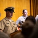 A senior U.S. Navy Chief Petty Officer in Service Khaki uniform is speaking at a podium during what appears to be a formal military ceremony or event. Multiple service members in lighter utility uniforms are visible in the background. The setting appears to be an indoor auditorium or ceremonial hall with wood-paneled walls.