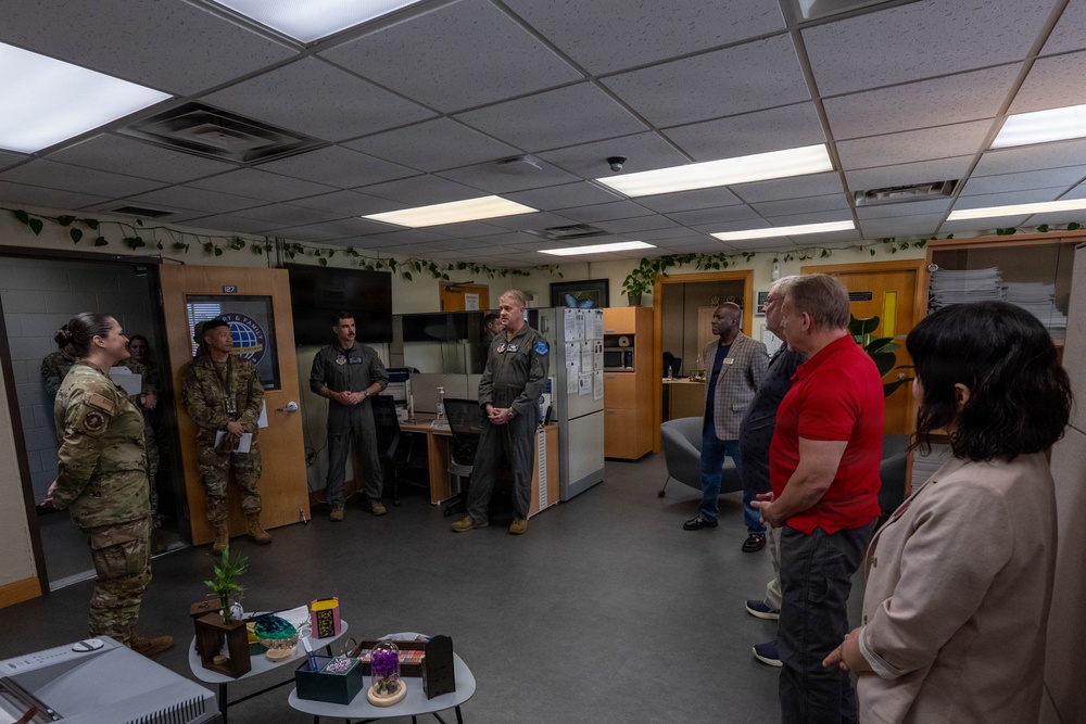 A group of military personnel in uniform and civilians are gathered in what appears to be a military office or support center, arranged in a semicircle as if during a briefing or tour. The room contains office furniture, a large monitor, and decorative plants along the walls. A female service member on the left appears to be presenting or briefing the group.