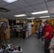 A group of military personnel in uniform and civilians are gathered in what appears to be a military office or support center, arranged in a semicircle as if during a briefing or tour. The room contains office furniture, a large monitor, and decorative plants along the walls. A female service member on the left appears to be presenting or briefing the group.