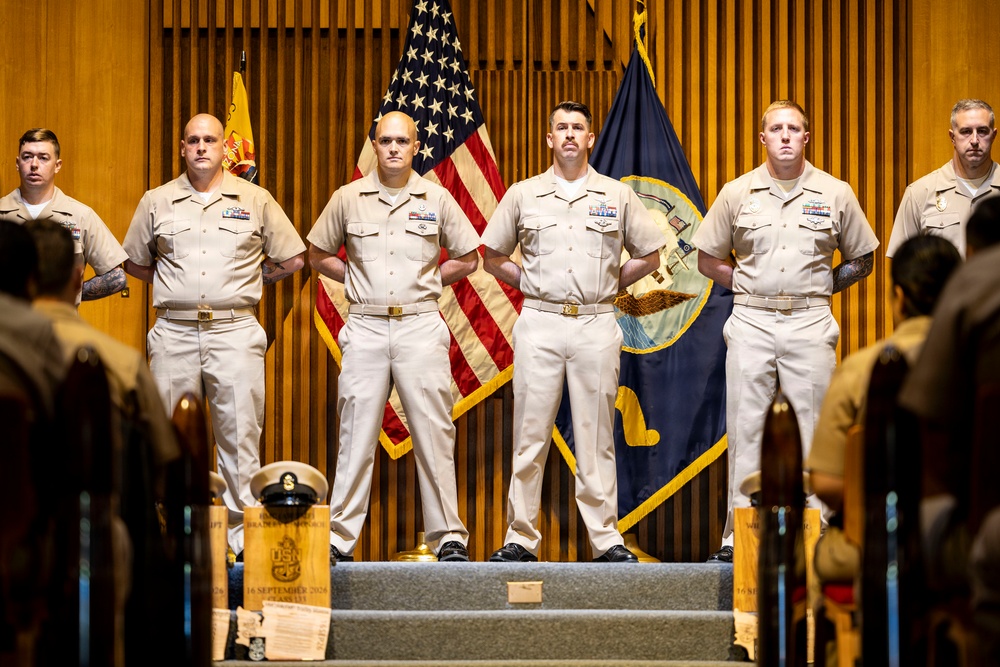A formal U.S. Navy ceremony featuring six sailors in Service Khaki uniforms standing at attention on a raised platform. The backdrop includes the American flag and a U.S. Navy flag against a wood-paneled wall. Engraved wooden plaques and a cover (hat) are visible in the foreground, suggesting a promotion or chief petty officer pinning ceremony.