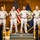 A formal U.S. Navy ceremony featuring six sailors in Service Khaki uniforms standing at attention on a raised platform. The backdrop includes the American flag and a U.S. Navy flag against a wood-paneled wall. Engraved wooden plaques and a cover (hat) are visible in the foreground, suggesting a promotion or chief petty officer pinning ceremony.