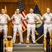 A formal U.S. Navy ceremony featuring six sailors in Service Khaki uniforms standing at attention on a raised platform. The backdrop includes the American flag and a U.S. Navy flag against a wood-paneled wall. Engraved wooden plaques and a cover (hat) are visible in the foreground, suggesting a promotion or chief petty officer pinning ceremony.
