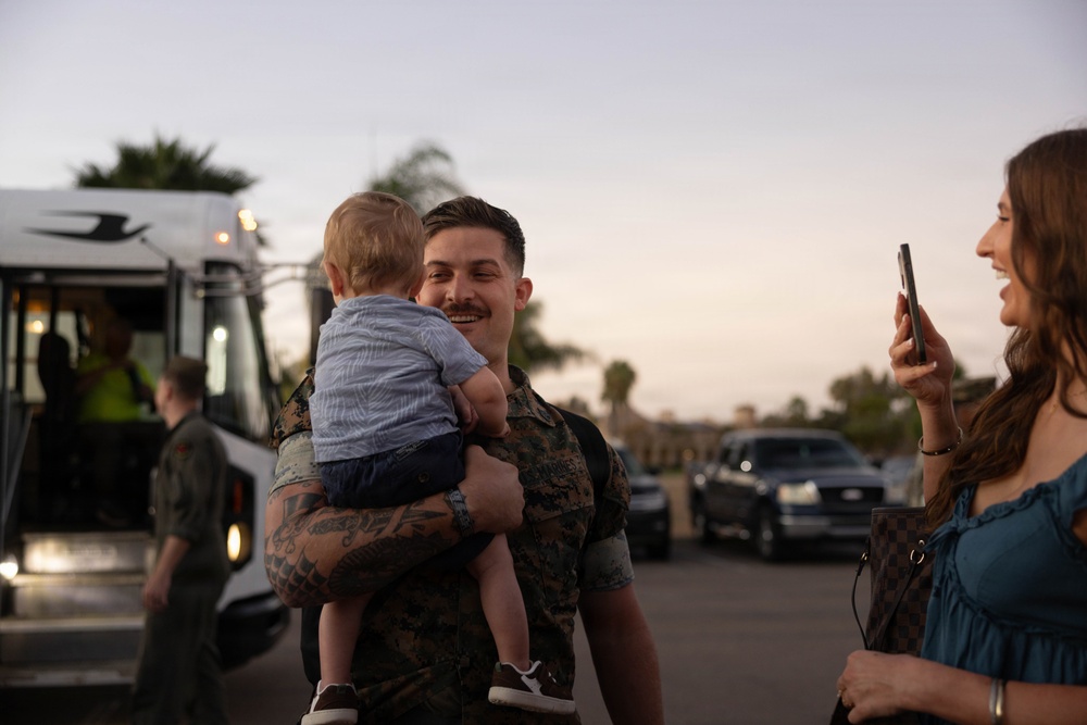 A service member in military camouflage uniform holds a young infant while smiling, as a woman photographs the moment on a smartphone. The scene appears to take place in a parking area near a bus, suggesting a military homecoming or reunion event. The warm, candid atmosphere and emotional expressions indicate a joyful family reunion.