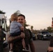 A service member in military camouflage uniform holds a young infant while smiling, as a woman photographs the moment on a smartphone. The scene appears to take place in a parking area near a bus, suggesting a military homecoming or reunion event. The warm, candid atmosphere and emotional expressions indicate a joyful family reunion.