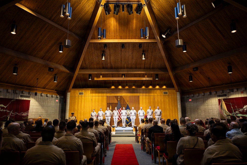 A formal military ceremony is taking place inside a chapel or auditorium with a distinctive A-frame wooden ceiling. A group of uniformed personnel in white dress uniforms stand on a raised stage, flanked by American and military branch flags, while a large audience of military personnel in tan/khaki uniforms and some civilians observe from wooden pew-style seating. A red carpet runs down the central aisle toward the stage.