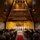 A formal military ceremony is taking place inside a chapel or auditorium with a distinctive A-frame wooden ceiling. A group of uniformed personnel in white dress uniforms stand on a raised stage, flanked by American and military branch flags, while a large audience of military personnel in tan/khaki uniforms and some civilians observe from wooden pew-style seating. A red carpet runs down the central aisle toward the stage.
