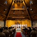 A formal military ceremony is taking place inside a chapel or auditorium with a distinctive A-frame wooden ceiling. A group of uniformed personnel in white dress uniforms stand on a raised stage, flanked by American and military branch flags, while a large audience of military personnel in tan/khaki uniforms and some civilians observe from wooden pew-style seating. A red carpet runs down the central aisle toward the stage.