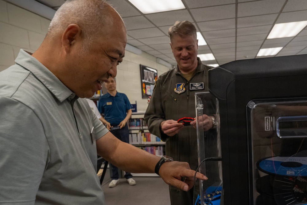 Two men are examining what appears to be a 3D printer in what looks like a library or resource room setting. One man in civilian clothes is interacting with the machine's controls, while a military officer in an olive flight suit observes and holds a small red object. A third person in a blue shirt is visible in the background near bookshelves.