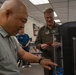Two men are examining what appears to be a 3D printer in what looks like a library or resource room setting. One man in civilian clothes is interacting with the machine's controls, while a military officer in an olive flight suit observes and holds a small red object. A third person in a blue shirt is visible in the background near bookshelves.