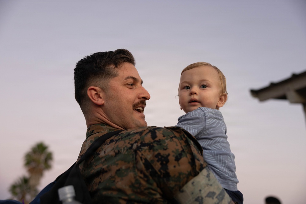 A man wearing U.S. Marine Corps MARPAT camouflage uniform is holding a young infant, approximately 6-12 months old, at dusk or dawn. The man is smiling and looking at the baby, who is gazing outward toward the camera. The setting appears to be outdoors with a palm tree and a structure visible in the background.