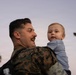 A man wearing U.S. Marine Corps MARPAT camouflage uniform is holding a young infant, approximately 6-12 months old, at dusk or dawn. The man is smiling and looking at the baby, who is gazing outward toward the camera. The setting appears to be outdoors with a palm tree and a structure visible in the background.