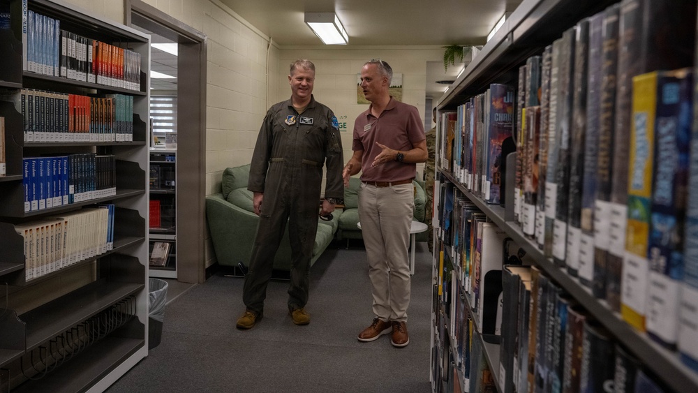 Two men are standing between bookshelves in what appears to be a military base library. One man is wearing an olive green military flight suit with patches, while the other is dressed in civilian attire (maroon polo shirt and khaki pants). They appear to be engaged in conversation while touring or reviewing the library facility.