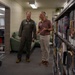 Two men are standing between bookshelves in what appears to be a military base library. One man is wearing an olive green military flight suit with patches, while the other is dressed in civilian attire (maroon polo shirt and khaki pants). They appear to be engaged in conversation while touring or reviewing the library facility.