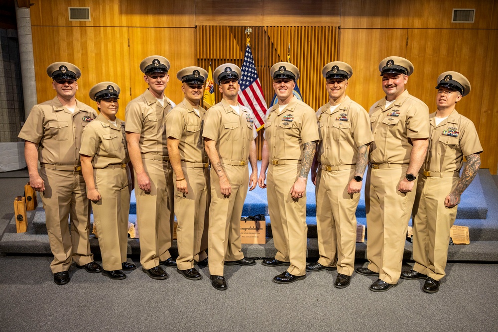 A group of nine U.S. Navy personnel in khaki service uniforms pose together in a formal indoor setting, likely during a promotion or recognition ceremony. They are standing in front of an American flag and what appears to be a Navy flag, with a wooden paneled backdrop. A partially visible sign in the foreground appears to read 'Congratulations on your promotion.'