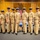 A group of nine U.S. Navy personnel in khaki service uniforms pose together in a formal indoor setting, likely during a promotion or recognition ceremony. They are standing in front of an American flag and what appears to be a Navy flag, with a wooden paneled backdrop. A partially visible sign in the foreground appears to read 'Congratulations on your promotion.'