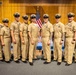 A group of nine U.S. Navy personnel in khaki service uniforms pose together in a formal indoor setting, likely during a promotion or recognition ceremony. They are standing in front of an American flag and what appears to be a Navy flag, with a wooden paneled backdrop. A partially visible sign in the foreground appears to read 'Congratulations on your promotion.'