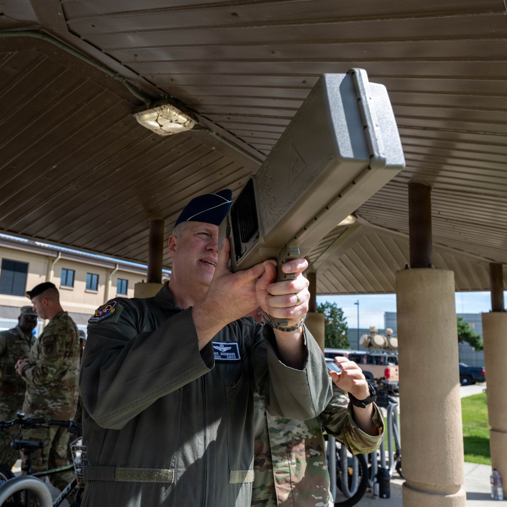 A U.S. Air Force officer in a flight suit is aiming or examining a large handheld counter-drone (C-UAS) device under a covered outdoor area on what appears to be a military installation. A second person in OCP (Operational Camouflage Pattern) uniform assists or observes nearby. Additional military personnel are visible in the background.