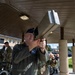 A U.S. Air Force officer in a flight suit is aiming or examining a large handheld counter-drone (C-UAS) device under a covered outdoor area on what appears to be a military installation. A second person in OCP (Operational Camouflage Pattern) uniform assists or observes nearby. Additional military personnel are visible in the background.