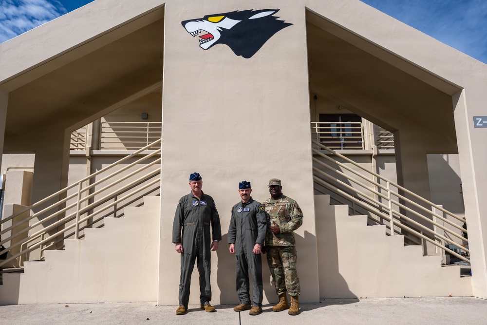 Three military personnel are posing together in front of a tan/beige military building featuring a wolf mascot logo painted on the facade. Two individuals are wearing olive green flight suits with Air Force-style caps, while the third is wearing Army Combat Uniform (ACU) in MultiCam pattern. The building has symmetrical staircases on both sides and appears to be a military installation facility.