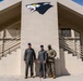 Three military personnel are posing together in front of a tan/beige military building featuring a wolf mascot logo painted on the facade. Two individuals are wearing olive green flight suits with Air Force-style caps, while the third is wearing Army Combat Uniform (ACU) in MultiCam pattern. The building has symmetrical staircases on both sides and appears to be a military installation facility.