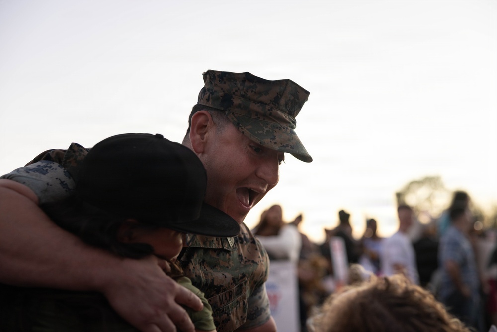 A U.S. Marine in digital camouflage uniform and cover (cap) is joyfully embracing a child, smiling broadly. The scene appears to be a military homecoming or reunion event, with a crowd of people visible in the blurred background. The emotional moment is captured at what appears to be dusk or dawn based on the soft, low light.