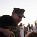 A U.S. Marine in digital camouflage uniform and cover (cap) is joyfully embracing a child, smiling broadly. The scene appears to be a military homecoming or reunion event, with a crowd of people visible in the blurred background. The emotional moment is captured at what appears to be dusk or dawn based on the soft, low light.