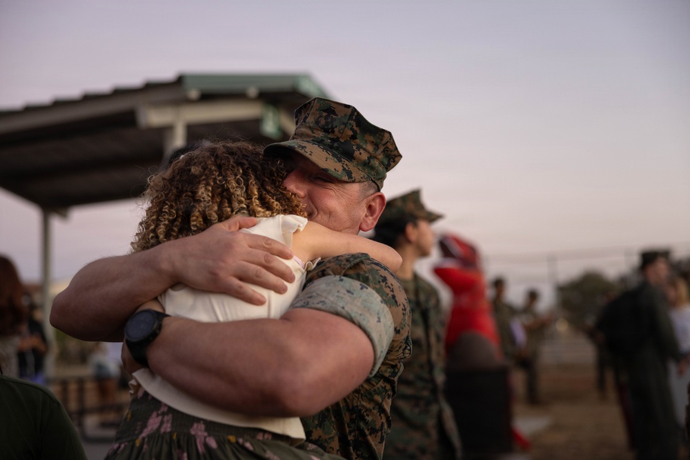 A military service member in U.S. Marine Corps camouflage uniform and cover (cap) embraces a civilian woman in an emotional hug. The setting appears to be an outdoor military base gathering, likely a homecoming or farewell event, with other people visible in the blurred background. The warm, low-light atmosphere suggests the scene is taking place near dusk.