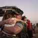 A military service member in U.S. Marine Corps camouflage uniform and cover (cap) embraces a civilian woman in an emotional hug. The setting appears to be an outdoor military base gathering, likely a homecoming or farewell event, with other people visible in the blurred background. The warm, low-light atmosphere suggests the scene is taking place near dusk.