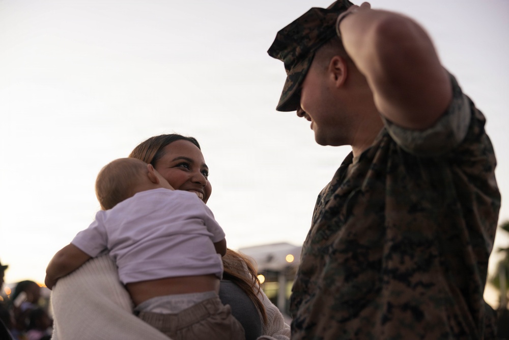 A U.S. Marine in digital camouflage (MARPAT) uniform and cover (cap) is facing a smiling woman who is holding an infant. The scene appears to be an emotional reunion, likely a military homecoming event, taking place at dusk or dawn outdoors. The woman's joyful expression and the intimate framing suggest a significant personal moment between the individuals.