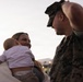 A U.S. Marine in digital camouflage (MARPAT) uniform and cover (cap) is facing a smiling woman who is holding an infant. The scene appears to be an emotional reunion, likely a military homecoming event, taking place at dusk or dawn outdoors. The woman's joyful expression and the intimate framing suggest a significant personal moment between the individuals.
