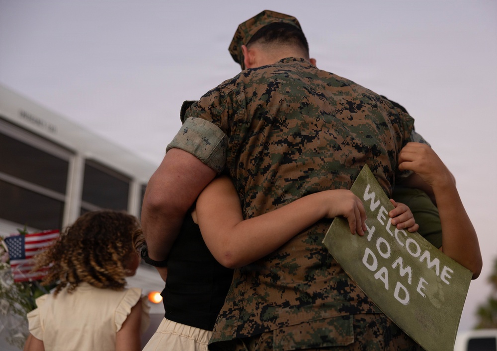 A military service member in MARPAT digital camouflage uniform is being embraced by a child holding a handmade 'Welcome Home Dad' sign. A second child with curly hair stands nearby, and an American flag is visible in the background. The scene depicts a military homecoming reunion, likely at a base or arrival point.