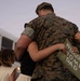 A military service member in MARPAT digital camouflage uniform is being embraced by a child holding a handmade 'Welcome Home Dad' sign. A second child with curly hair stands nearby, and an American flag is visible in the background. The scene depicts a military homecoming reunion, likely at a base or arrival point.